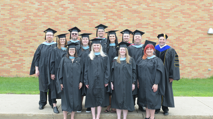 Group of Master's degree candidates posing in front of a brick wall in academic regalia on commencement day at Valley City State University
